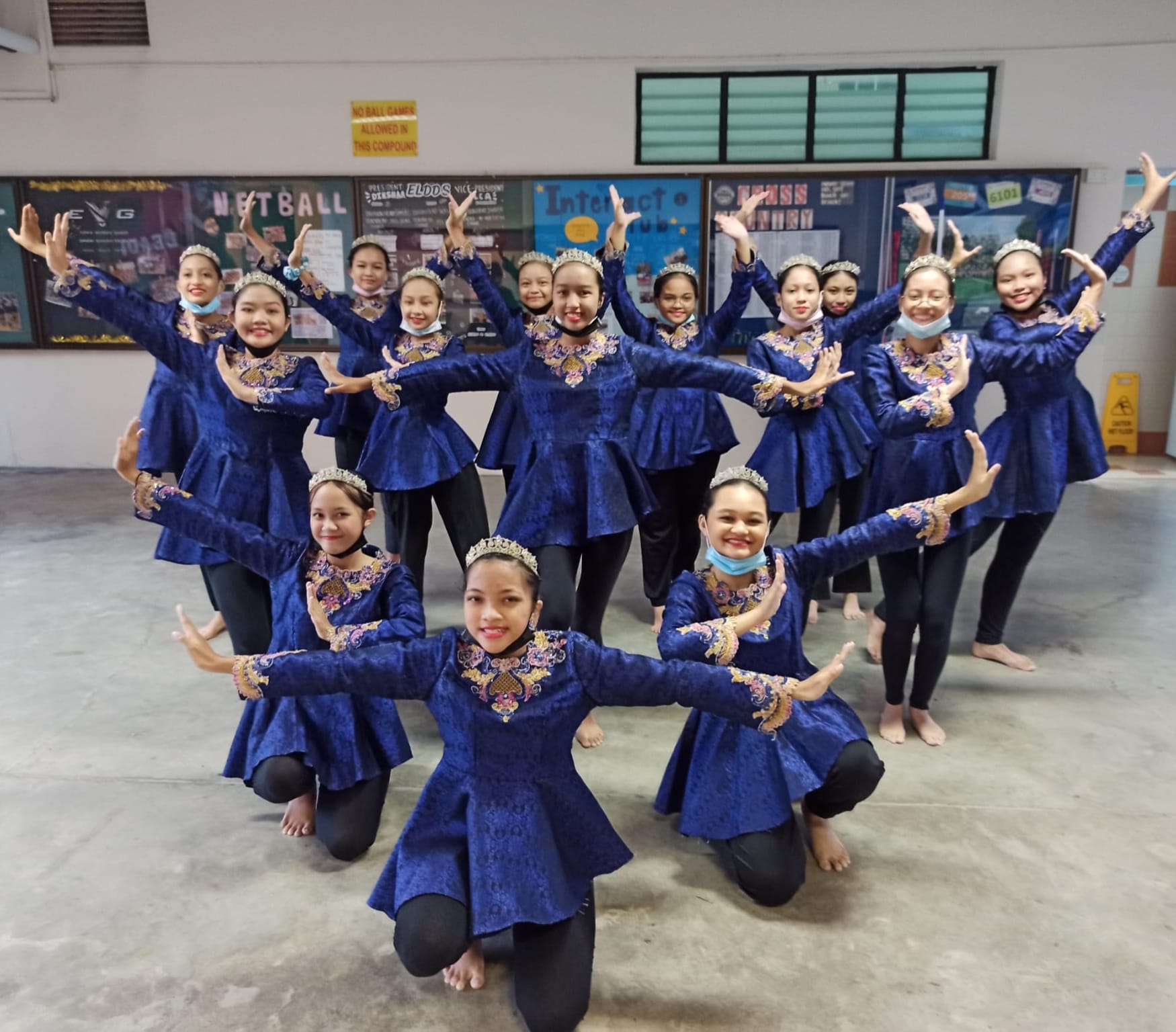 A group of young female dancers in matching blue embroidered costumes and tiaras pose together indoors.